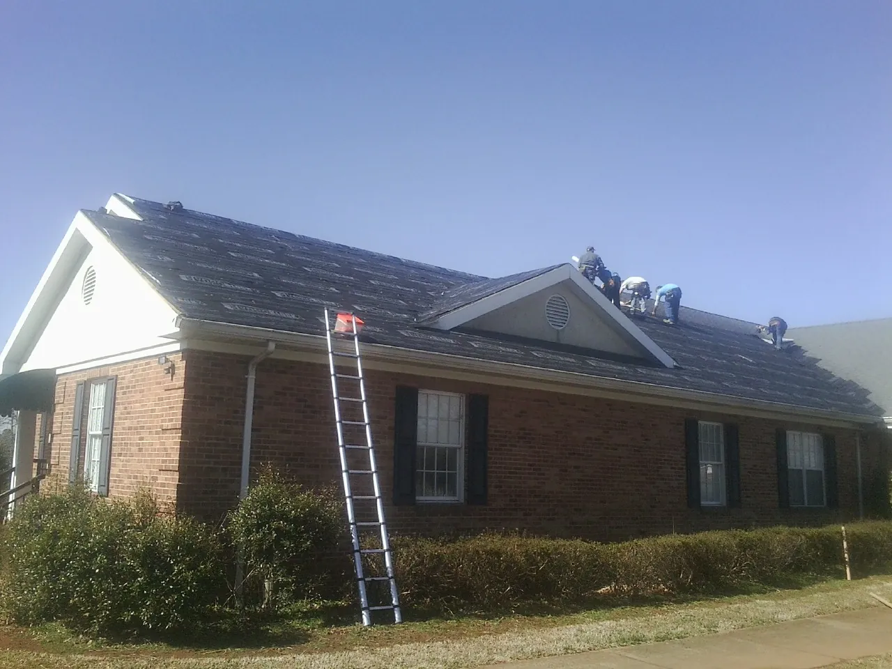 Crew tearing shingles off a brick ranch roof with a ladder set — Emergency Roof Repair work by our crew in Imlay City