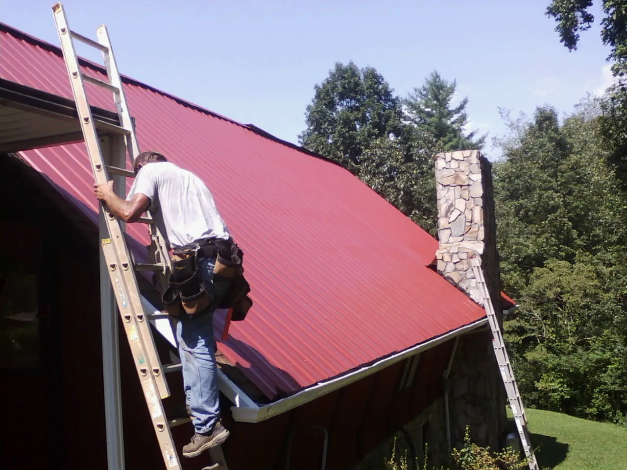 Roofer on a ladder beside a red metal roof and stone chimney — Soffit & Fascia Repair by Roofing Brothers in Imlay City